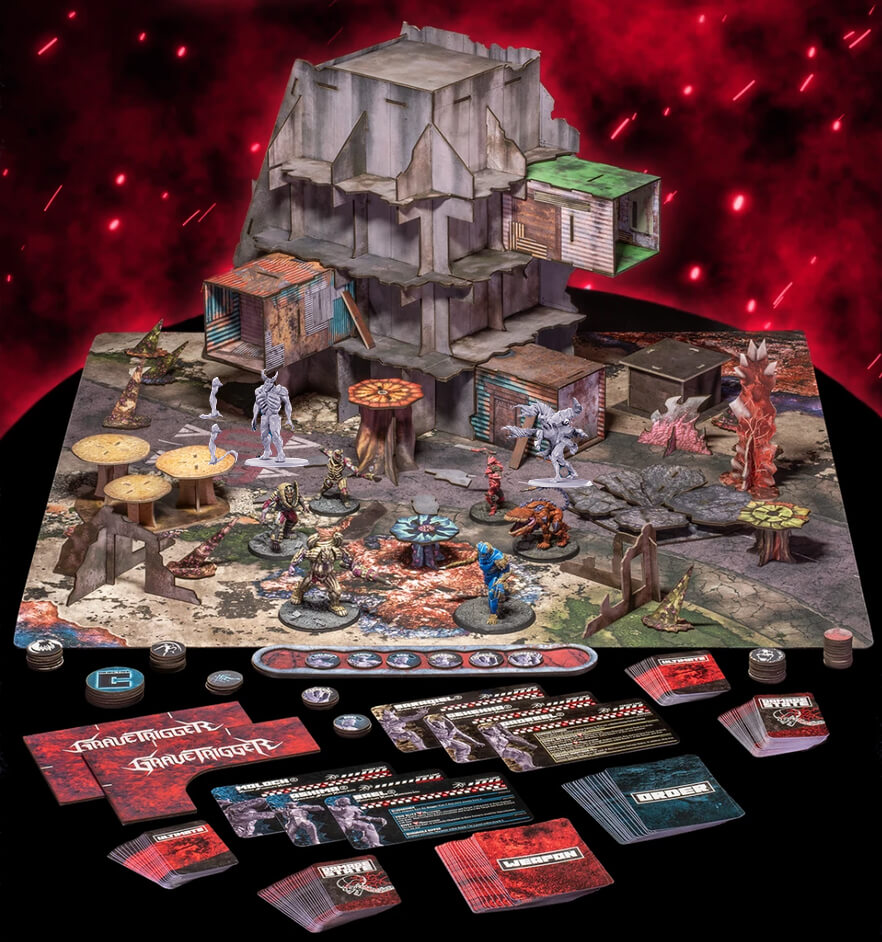 A tabletop display of a post-apocalyptic  tabletop gaming set, featuring a large detailed terrain mat with ruined buildings, tables, and monster miniatures, alongside numerous rulebooks, cards, dice, and tokens arranged on a black surface against a dramatic red glowing background.